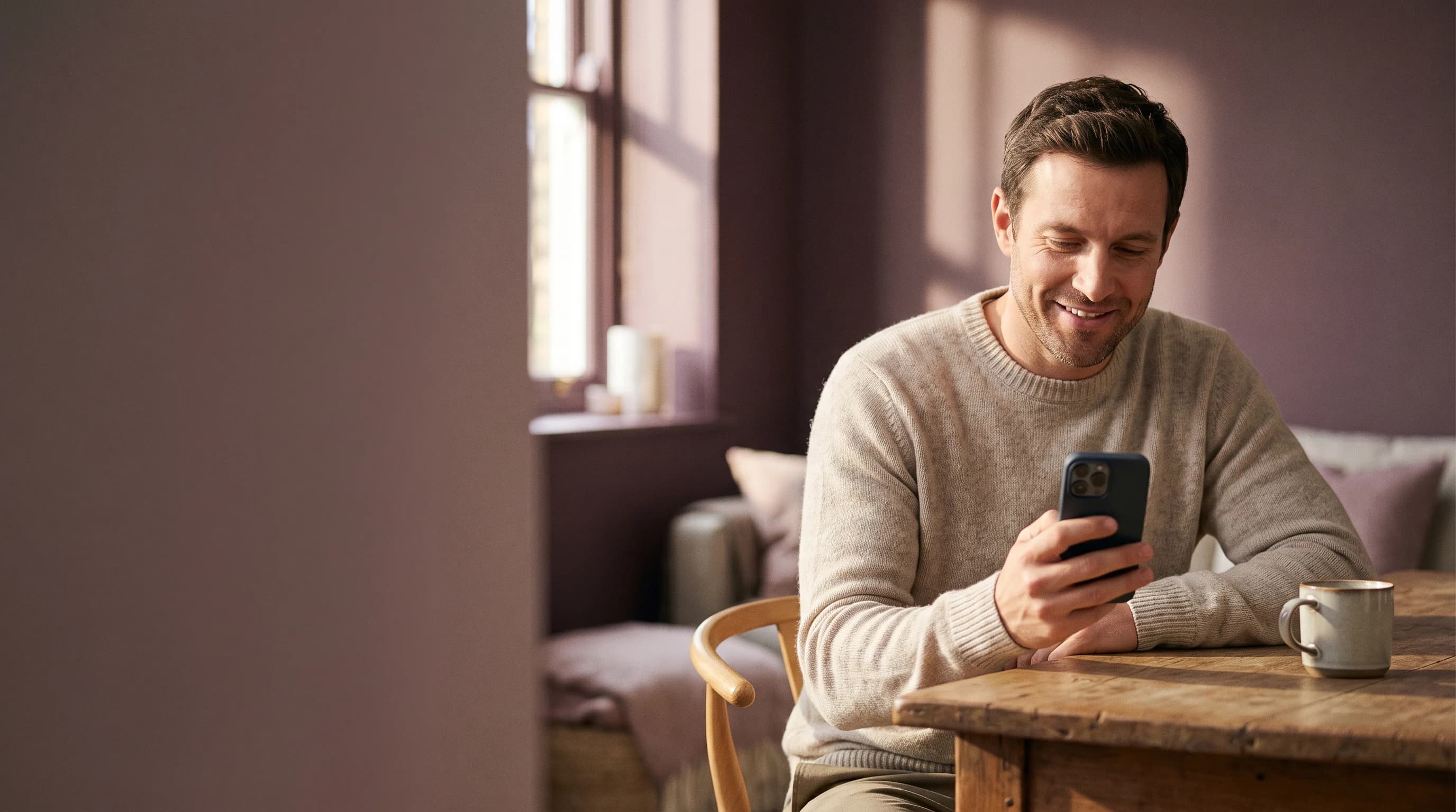 A smiling man in a cream jumper reads something on his phone at a wooden table by a window